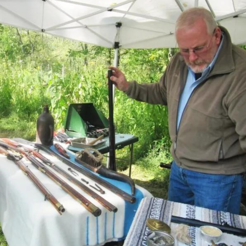 Image of Doug Plance, standing behind a table with several long rifles on it.