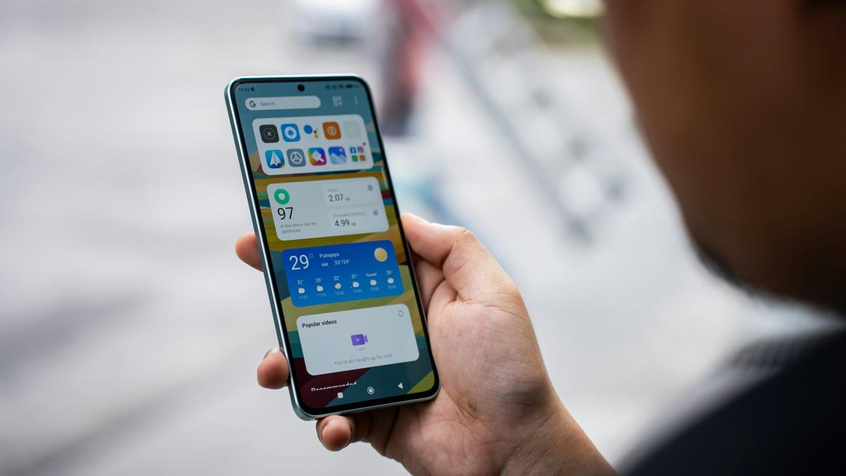 A person is holding a smartphone. It displays the home screen with various apps and the weather.